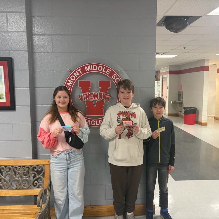 Three students standing in a hallway, one holding a box, in front of a wall with a school logo.