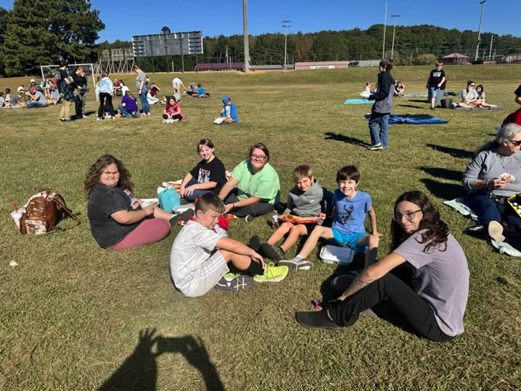 A group of people sit on a grass field in an open area with trees and a building.