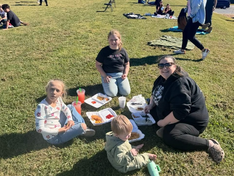 Family seated on grass eating, one adult holding a child, two adults wearing glasses, tray of food, drinks.