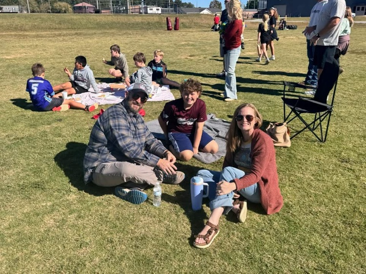 People sit on grass in a field. Some sit on blankets and others stand behind them. Some have backpacks.