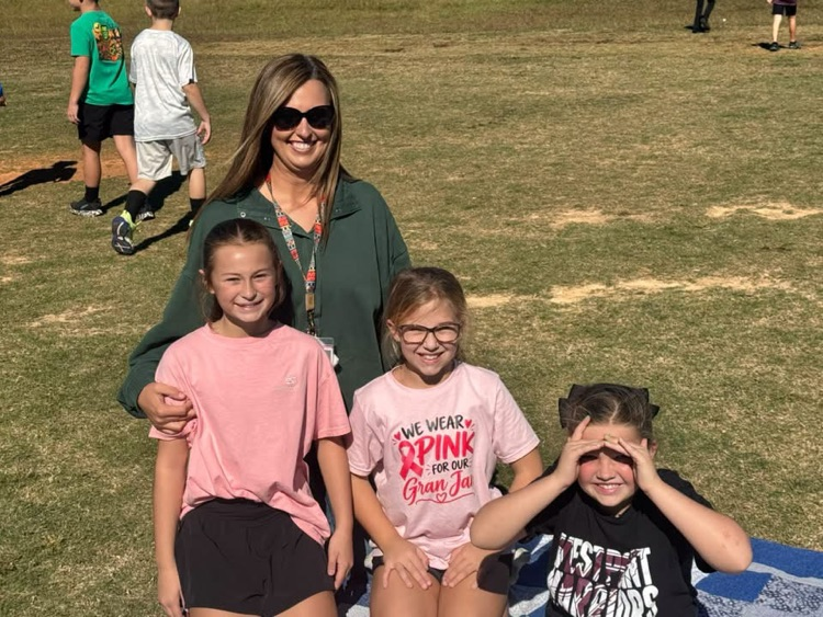 Adult woman and three children sitting on grass. Children wear pink shirts. One child wears glasses. One child has hand over eyes.