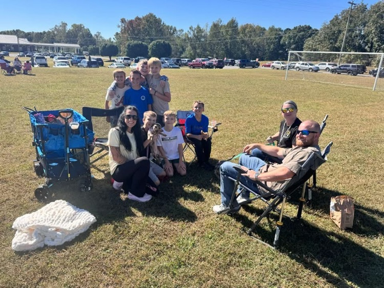 Family group sitting in folding chairs on a grassy field with a volleyball net in the background.