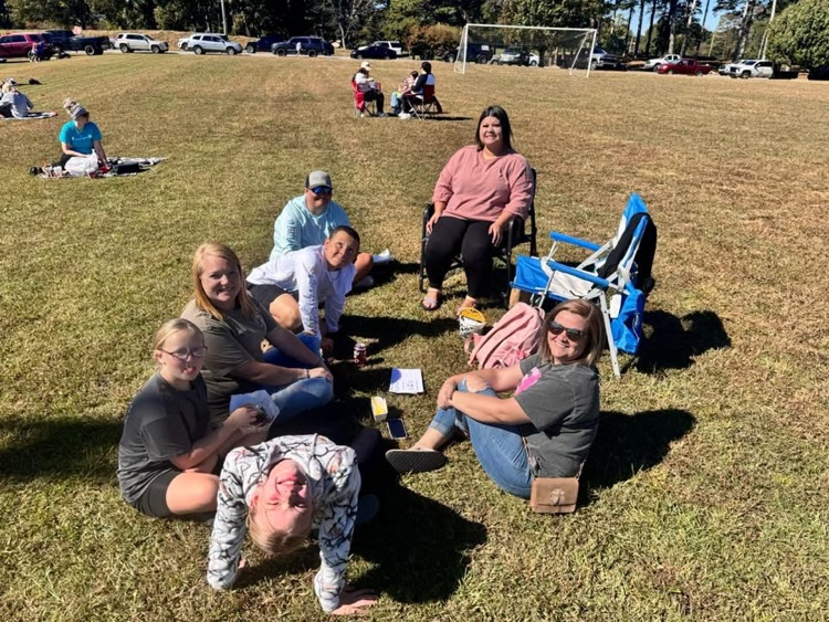 People sit and stand on grass in a park; some sit in chairs, one leans back on arms.