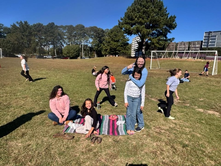 People gather in a grassy field. Some sit on a blanket while others stand, some wearing sneakers. Trees and buildings in the distance.