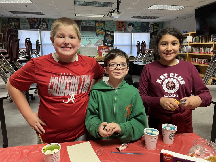 Three children stand behind a table with cups and bowls of paint. Two boys and one girl.
