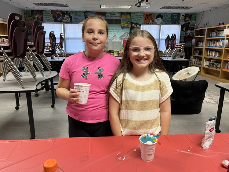 Two girls stand by a red table, holding cups. One holds a cup with a straw.