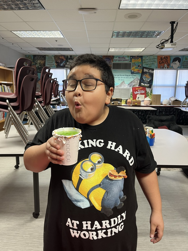 A boy holds a cup of green drink while standing in a classroom. The table has chairs and boxes.