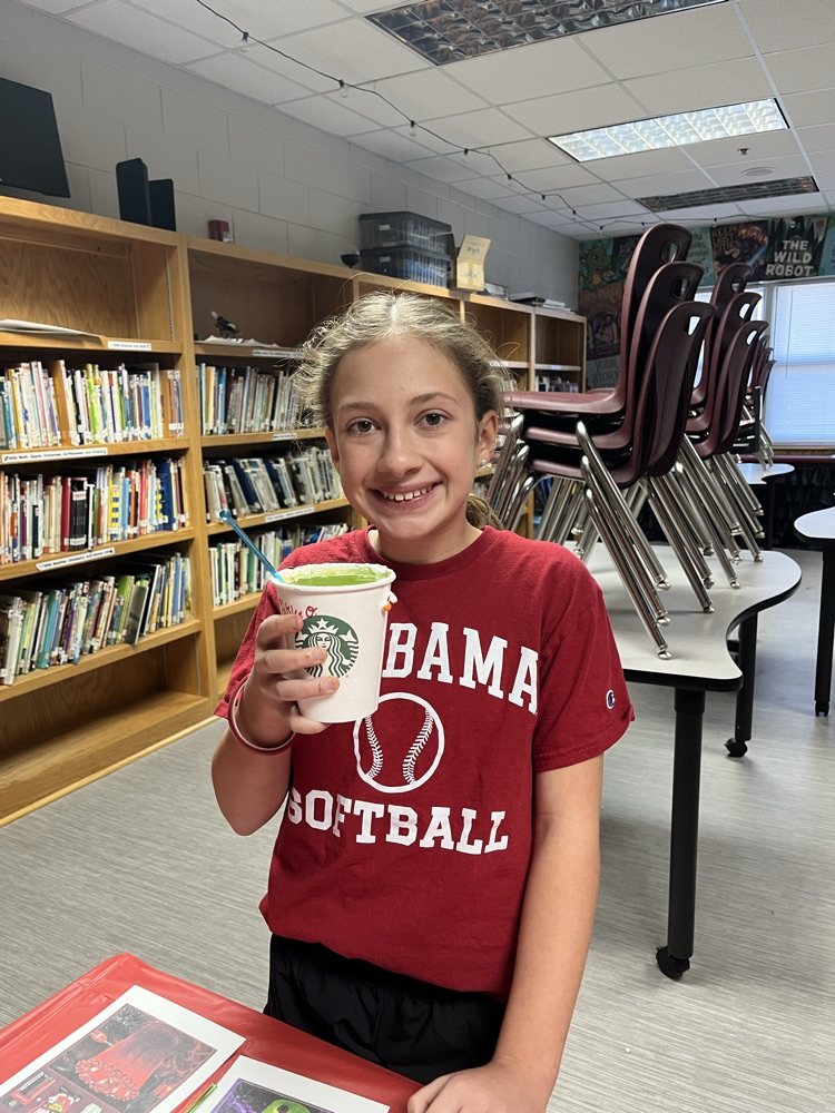 A girl in a red shirt holding a cup stands in front of shelves with books.