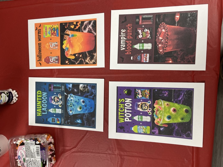 Four posters on a red table. One poster has a blue monster on it. Another poster has a green monster on it. There are candies in the foreground.
