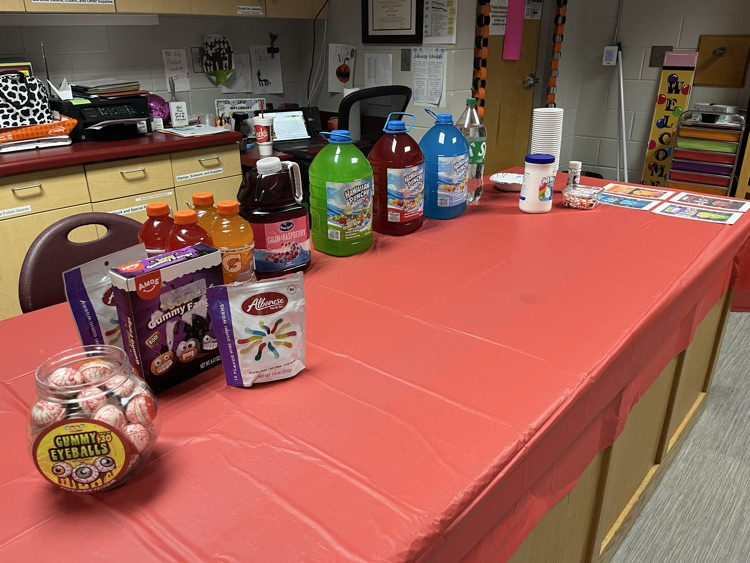 Table with red tablecloth, bottles of various drinks, bowl of balls, and boxes of snacks.