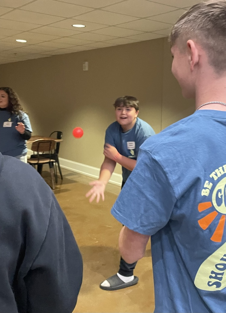 Four people in a room with chairs, one man throwing a ball at another man.