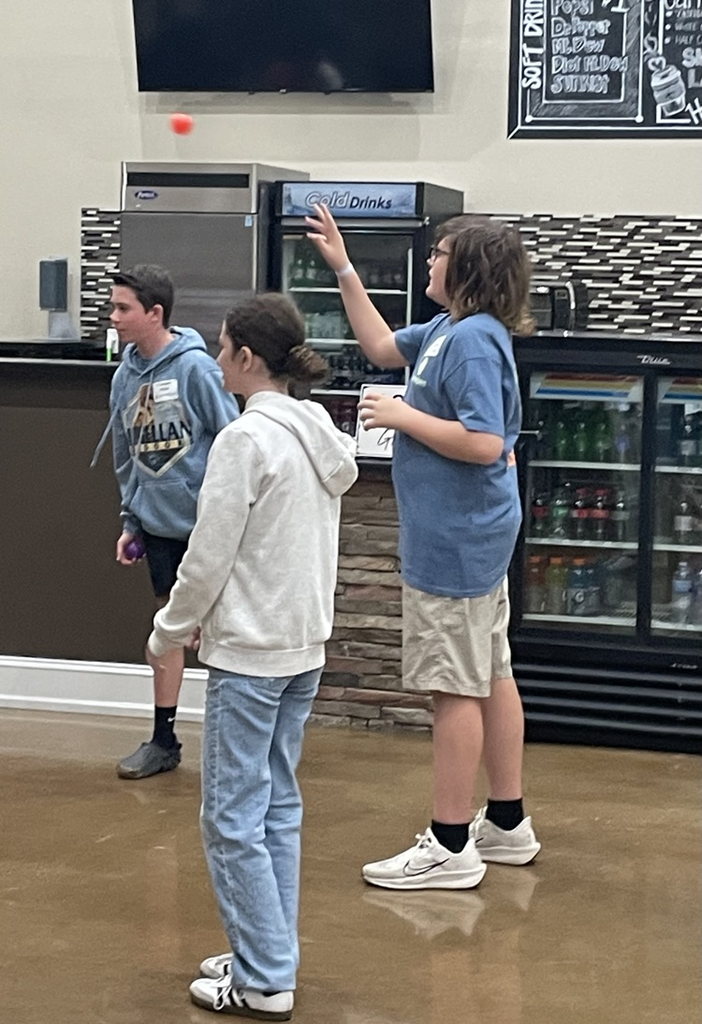 Three people in a cafeteria. One person raises their hand. Another person holds a small object.