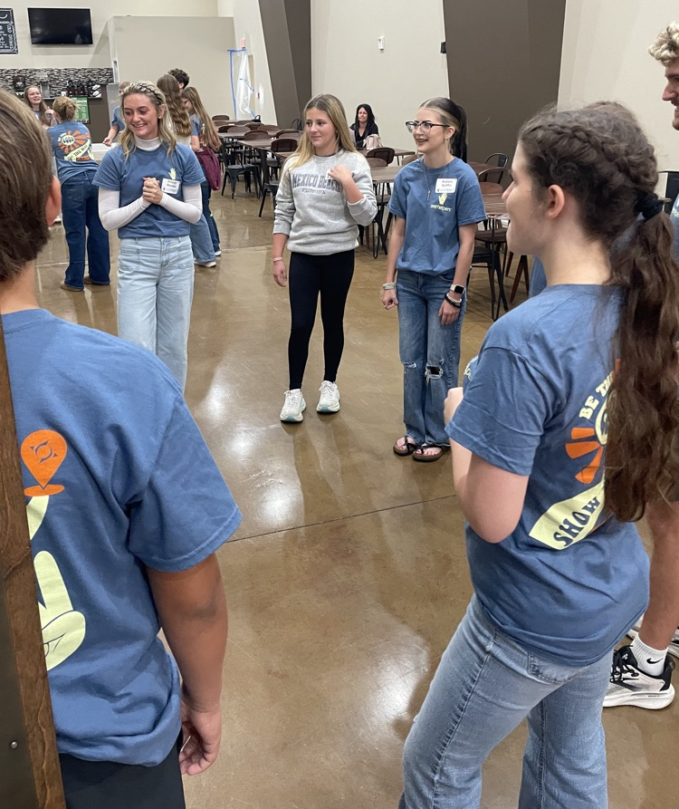 Five students in blue shirts, some with ponytails, stand in a hall. One faces away, two face the camera, and one speaks with hands.
