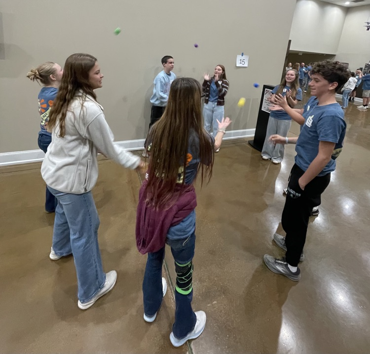 Several students play a game involving small yellow balls in a room with a white wall and concrete floor.