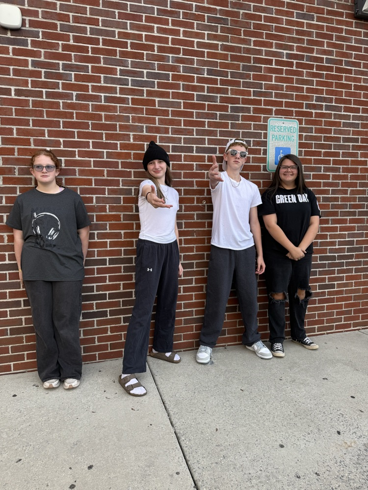 Four students stand in front of a red brick wall, each wearing casual clothes and shoes, with one gesturing.
