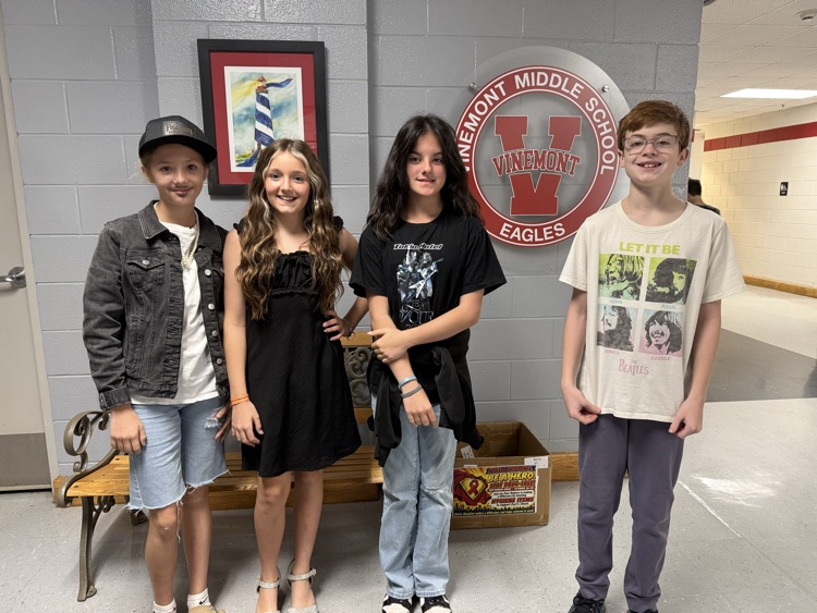 Four children standing in front of a bench, with a logo and framed art in the background.