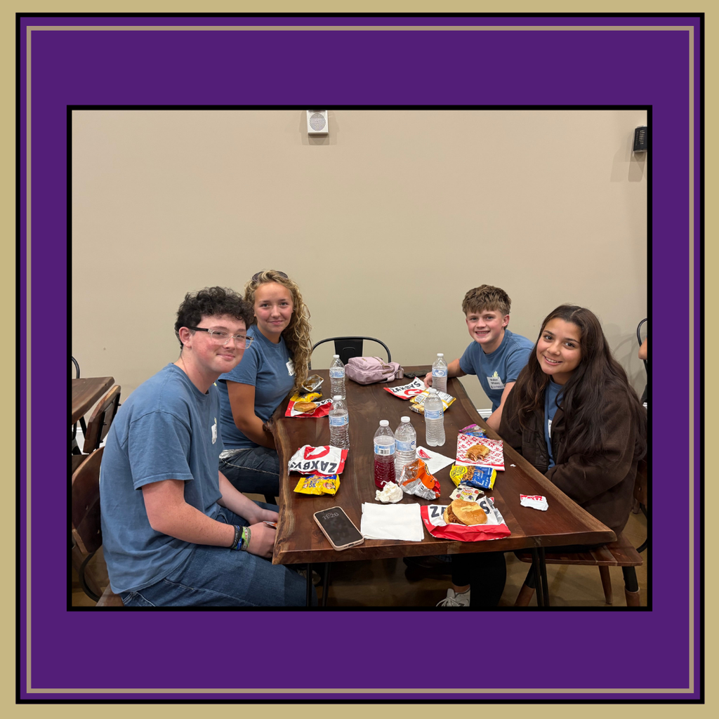 Five people sit around a table. They have snacks, drinks, and a phone on the table.