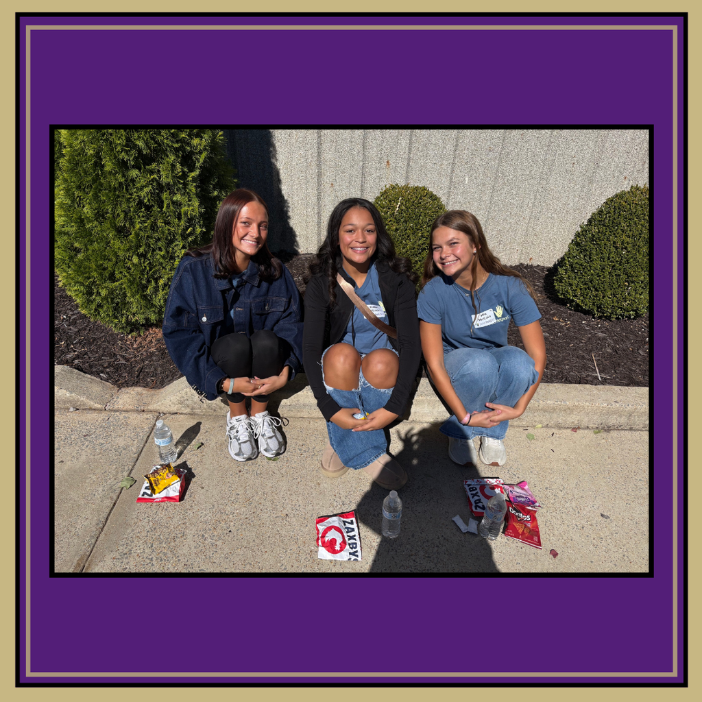 Three young women in blue shirts and jeans sit on a curb, posing for a photo.