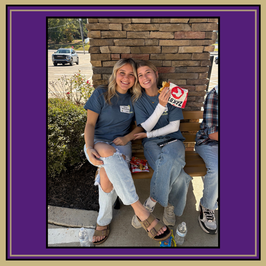 Two women sit on a bench, one holding food. A third person sits next to them. A brick wall and plants behind them.