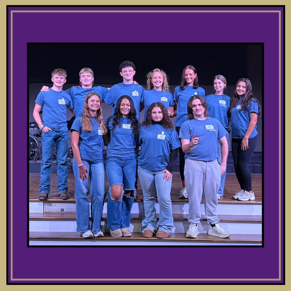 Several students stand on stage wearing blue shirts and jeans; two wear pants in different colors.