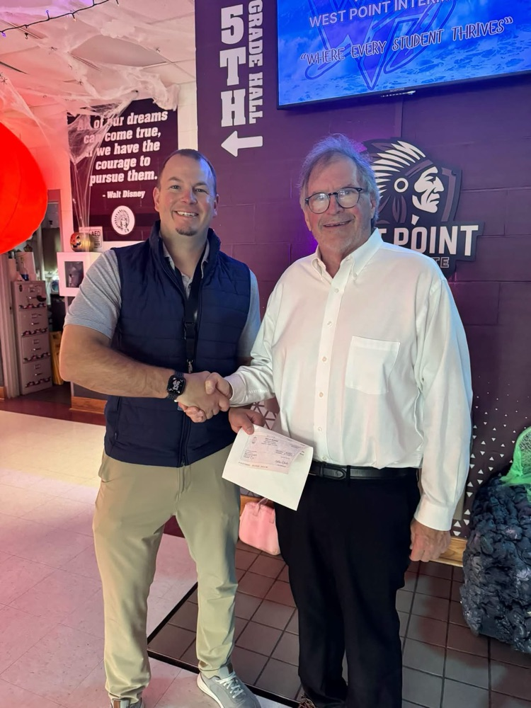 Two men stand and shake hands. A sign reading "5th Grade Hall" is in the background.