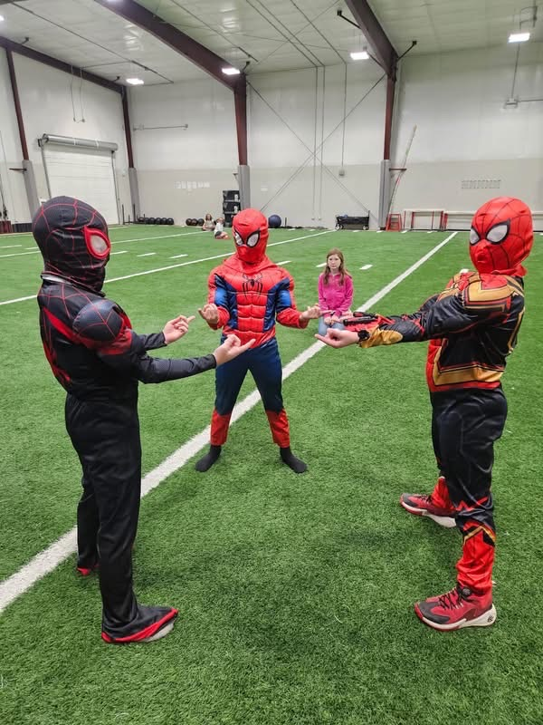 Three children in superhero costumes interact on a green field. A girl in pink stands apart.