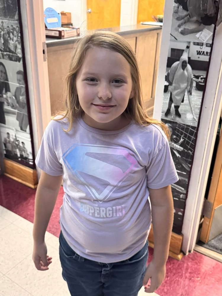 A young girl in a white shirt with a Superman logo smiles and poses for a photo.