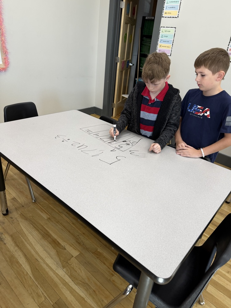 Two boys write on a white board with markers. One boy is wearing a blue shirt and the other a striped shirt.