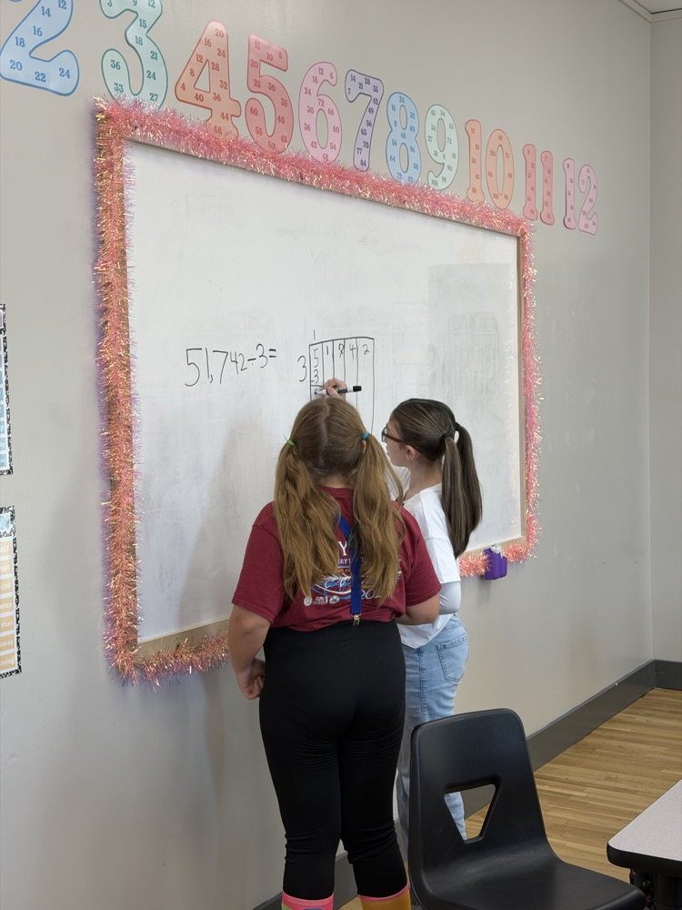 Two students standing in front of a whiteboard writing numbers. Whiteboard has colorful letters on the edge.