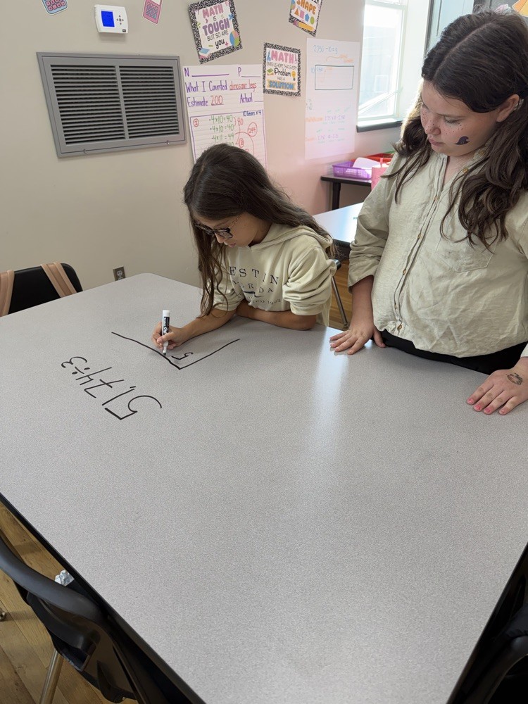 Two girls are writing on a long table. One girl is holding a pen and the other is looking.