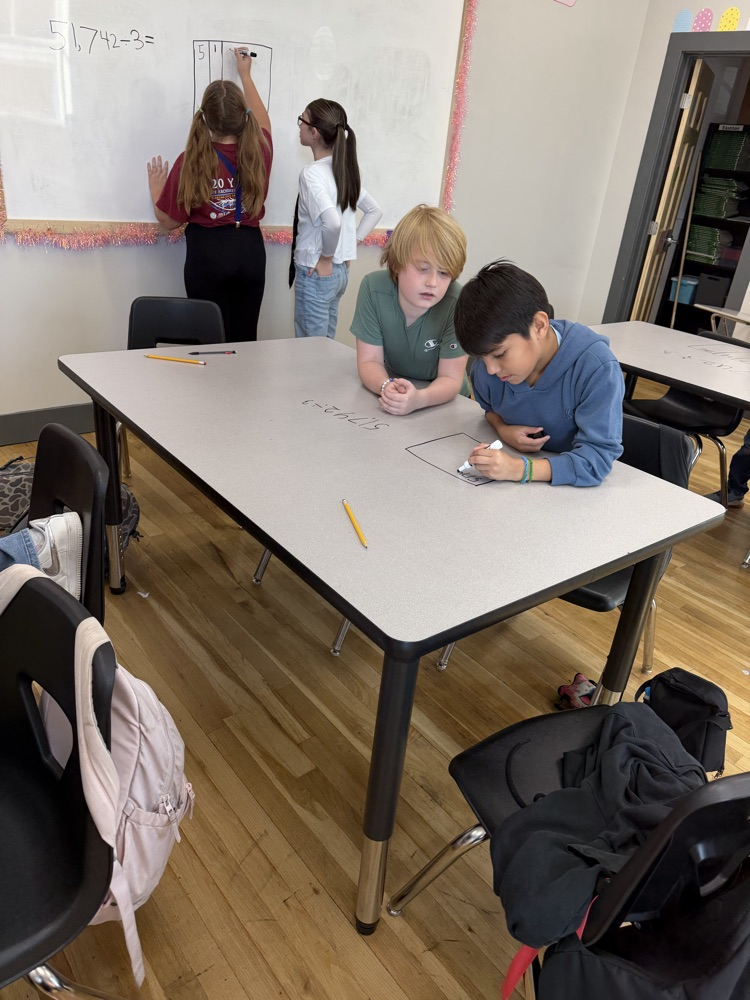 Students work at tables, writing on a whiteboard; a girl stands and writes while others sit.