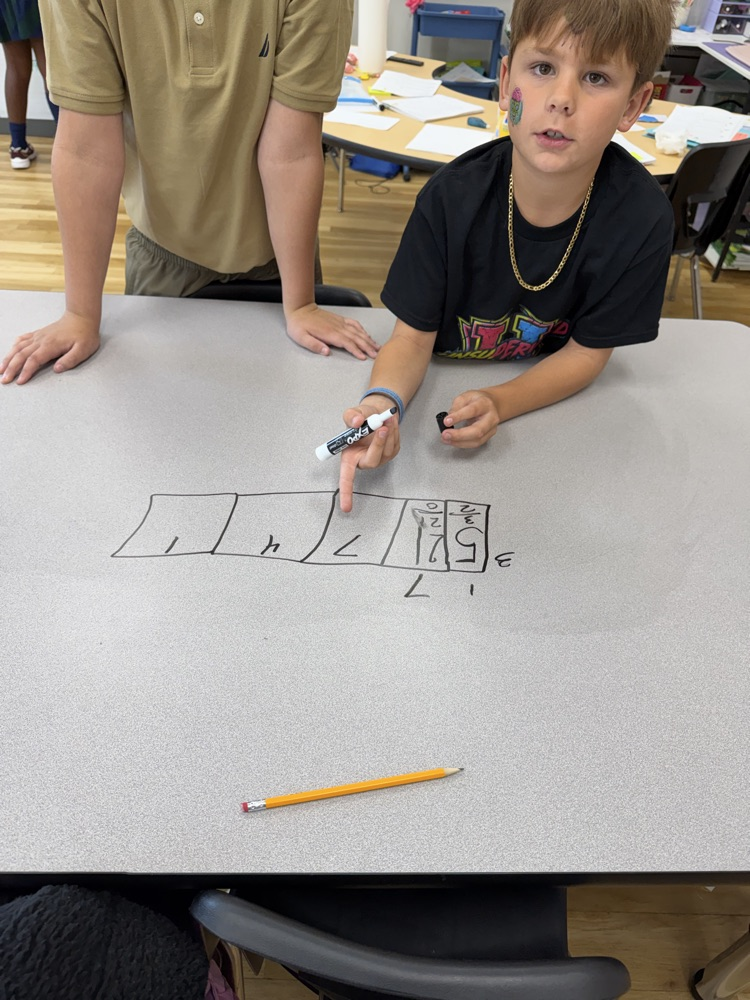 Two young boys seated at a table, one using a marker to draw a diagram.
