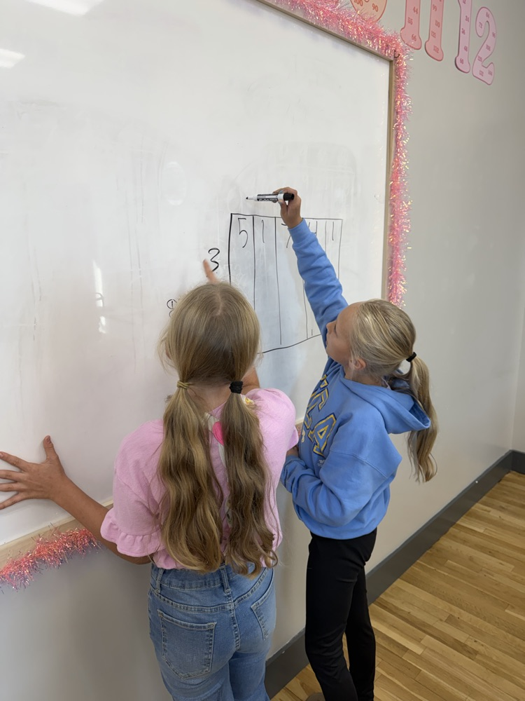 Two young girls writing on a whiteboard with a pink trim. One writing in blue marker, the other writing in black.