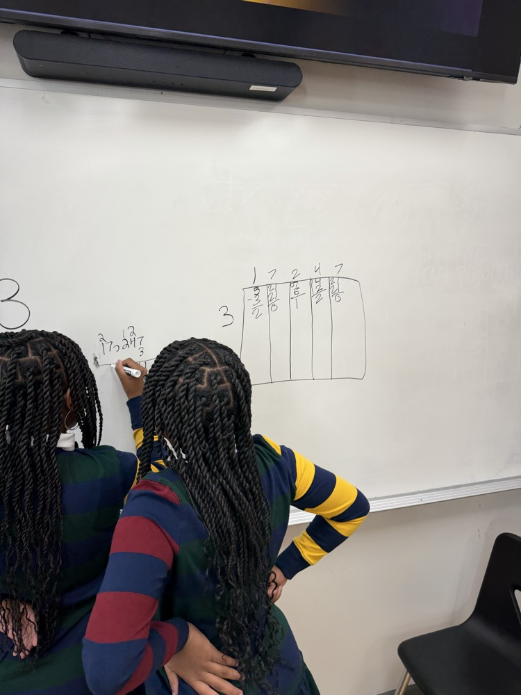 Two students write on a whiteboard with a number 3 and a chart. One student holds a marker.