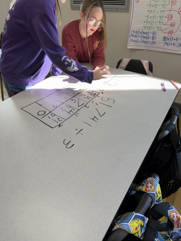 Two students are seated, one writing on a whiteboard with numbers and letters, the other wearing glasses.