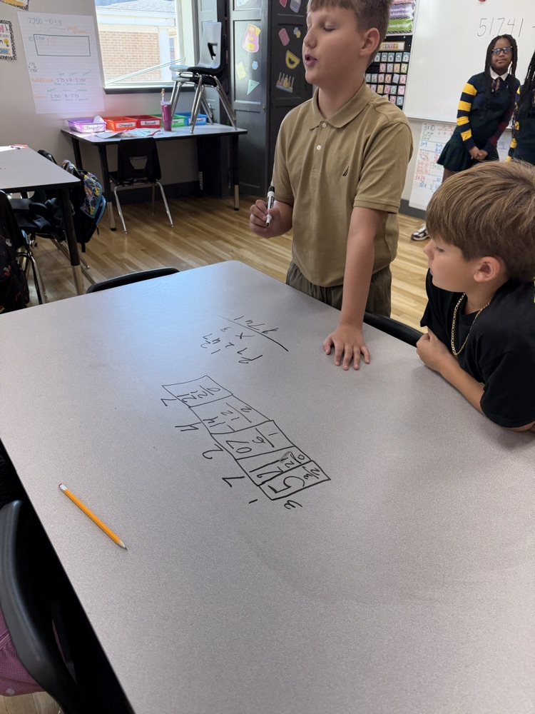 Two boys sit at a table with a whiteboard. A boy writes on the board while another boy watches.
