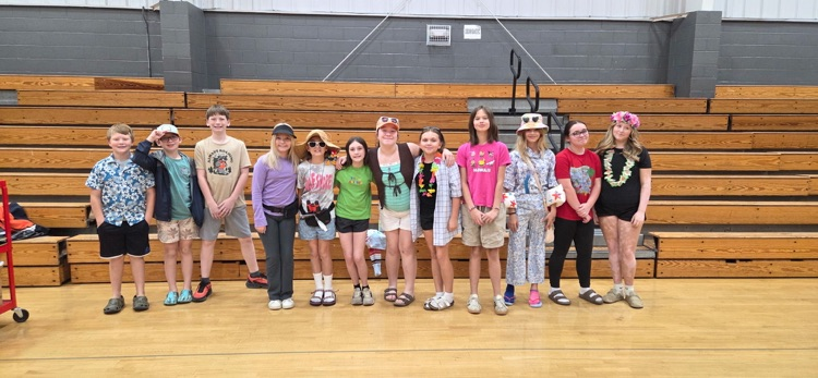 A group of children and young adults stands on a gym floor, wearing casual clothes, some with hats and necklaces.