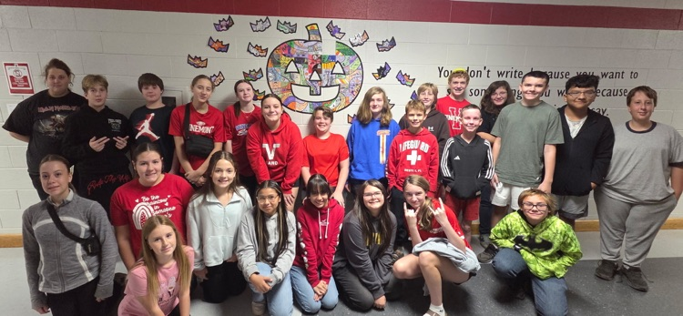 A group of students, some wearing red, crouch or stand against a white wall with a colorful circular design.