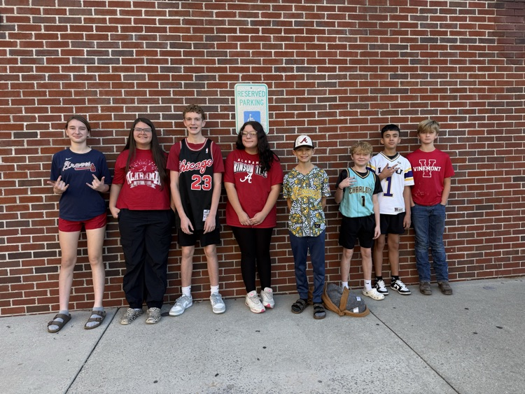 A group of young people pose outside a brick wall, some wearing hats and sneakers.