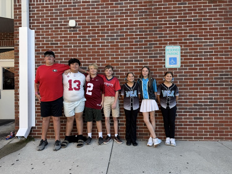 Seven students and one adult stand next to a brick building with a sign above the sidewalk.