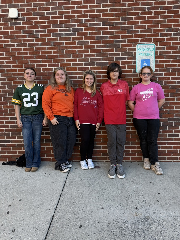 Five students stand next to a brick wall. They wear various colored shirts and pants. A sign on the wall reads Reserved Parking.