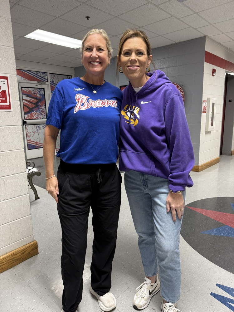 Two women pose for a photo in a school hallway. One wears a blue shirt and black pants. The other wears a purple hoodie and jeans.