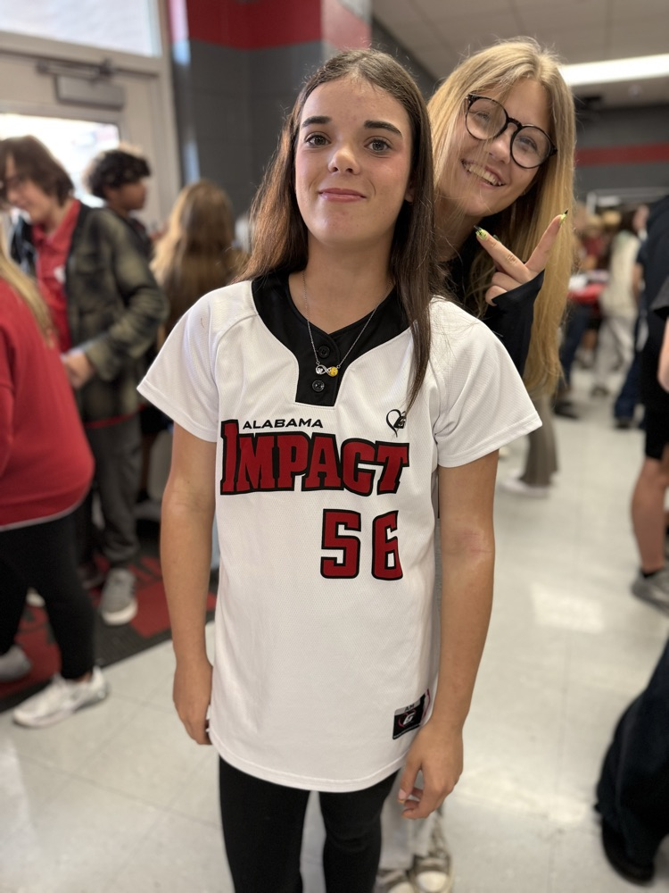A young girl in a white jersey with black trim stands next to an adult in a school hallway.
