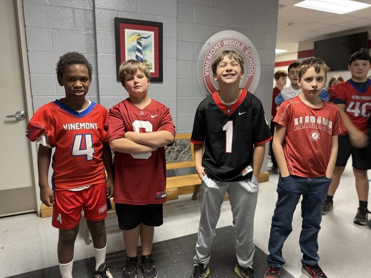 Four boys in sports uniforms stand in a corridor with a bench and framed picture behind them.