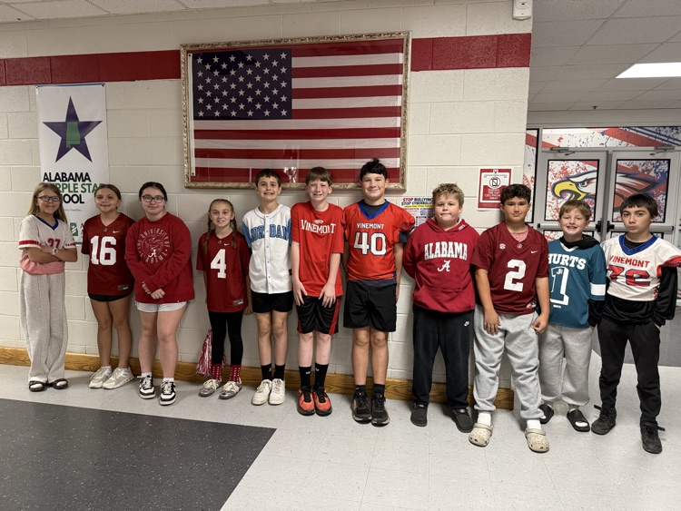 Several students in matching red shirts and numbered jerseys stand in a hallway, some with hands on hips, against a red wall with an American flag and banners.