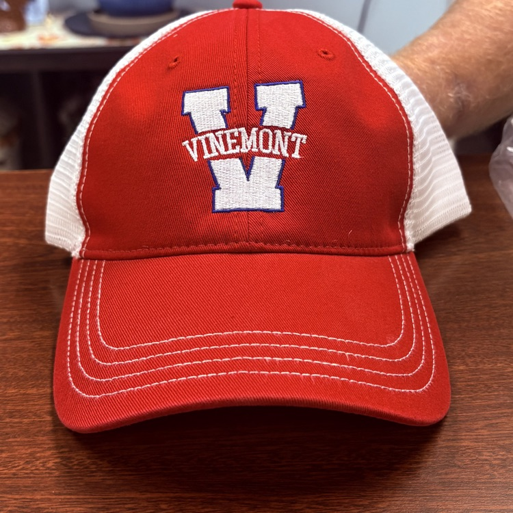 Red and white baseball cap with "Vinemont" and "V" on front sits on a wooden table.