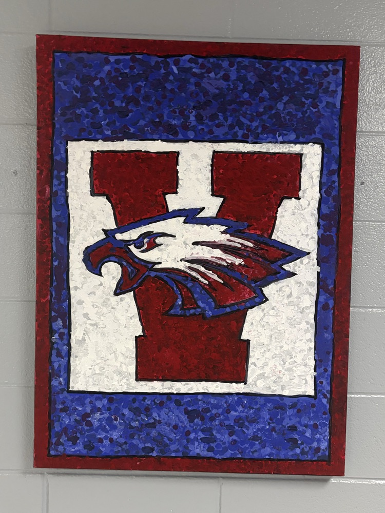 A framed art piece with a red and blue eagle over a white W is displayed on a white brick wall.