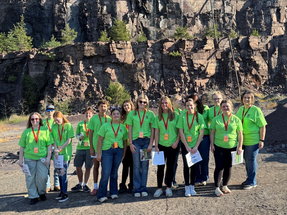 A group of Eastern View High School Future Farmers of America (FFA) students and staff stand together at Luck Stone Quarry in Culpeper. They are wearing bright green shirts and red lanyards, holding papers and smiling for the camera. Behind them is a dramatic rock wall from the quarry, with trees growing along the ledges.