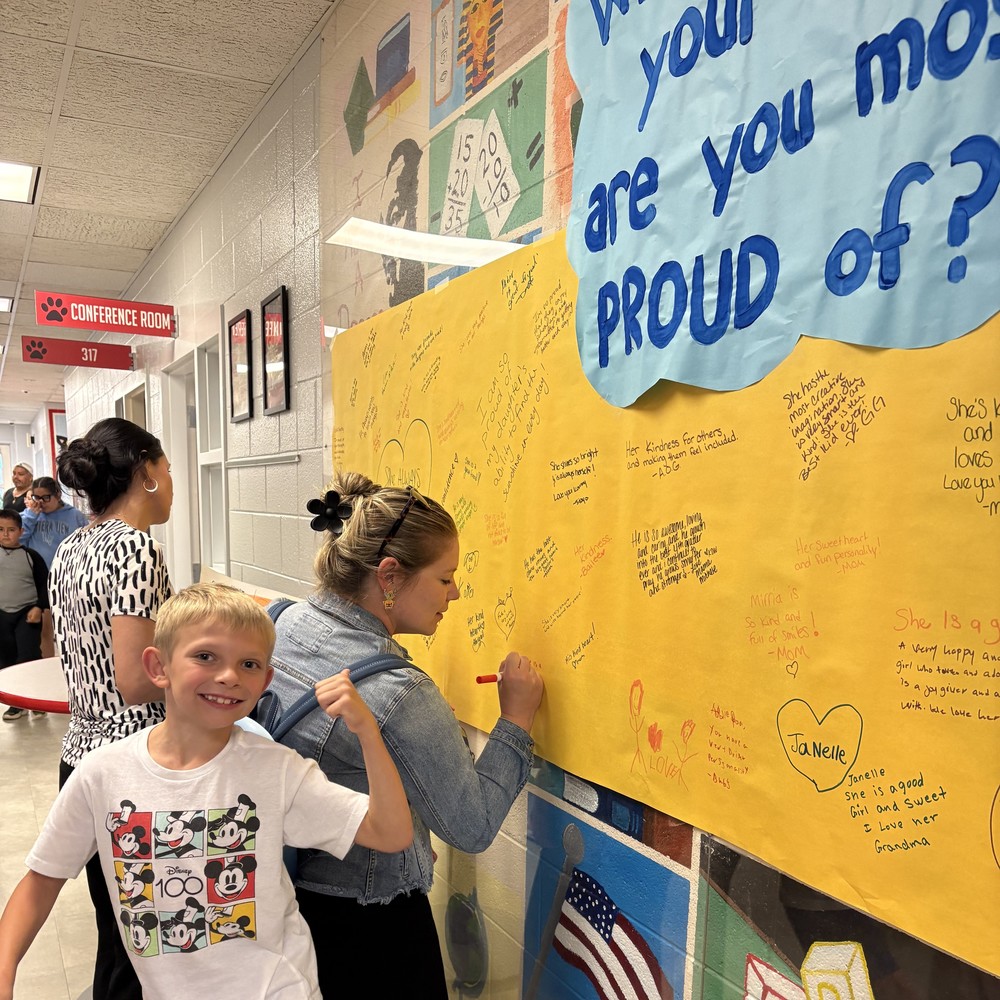 A young student smiles and flexes his arm in front of a large yellow “What are you most proud of your child?” wall at AG Richardson Elementary’s Back to School Night. Parents and family members are writing heartfelt notes on the wall, celebrating their children’s accomplishments.