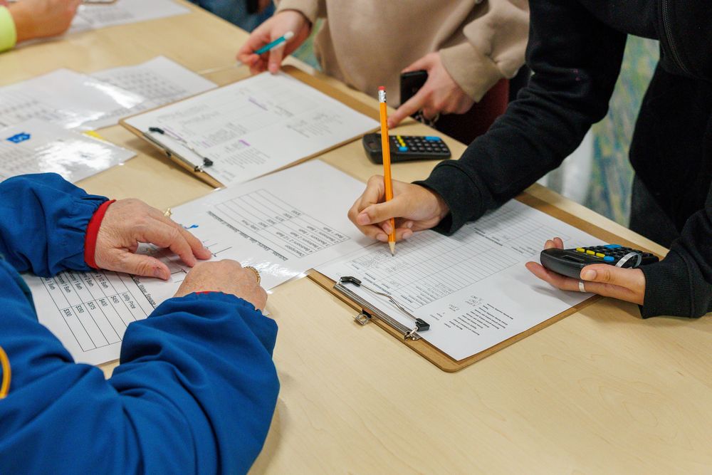 Close-up of several people working at a table with clipboards, calculators, and pencils. They appear to be filling out forms or worksheets, possibly engaging in a budgeting or data-entry activity. One person in a blue jacket points to a paper, while another writes on a chart with a pencil.